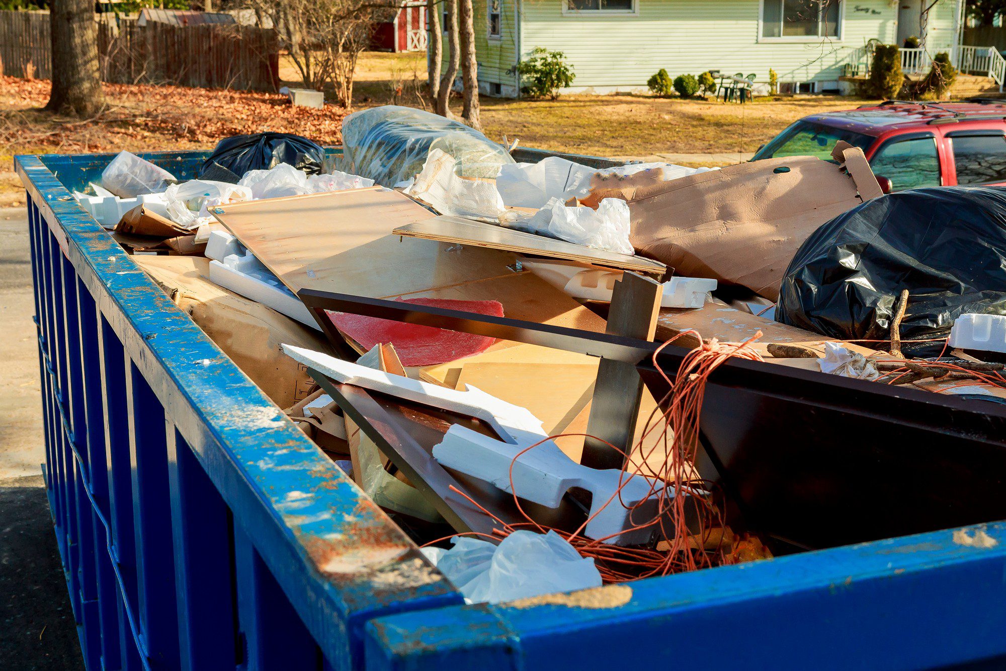 The image shows a large blue dumpster filled with various types of waste and discarded items. It appears to contain a mix of construction or renovation debris, such as pieces of wood, scraps of drywall, broken furniture parts, plastic bags, and other materials. Some items like polystyrene foam and wires can also be seen among the debris. The dumpster is located in an outdoor area with residential housing in the background, and there are cars parked nearby suggesting this could be near an flat complex or a residential street where some kind of construction or cleanup project is underway.