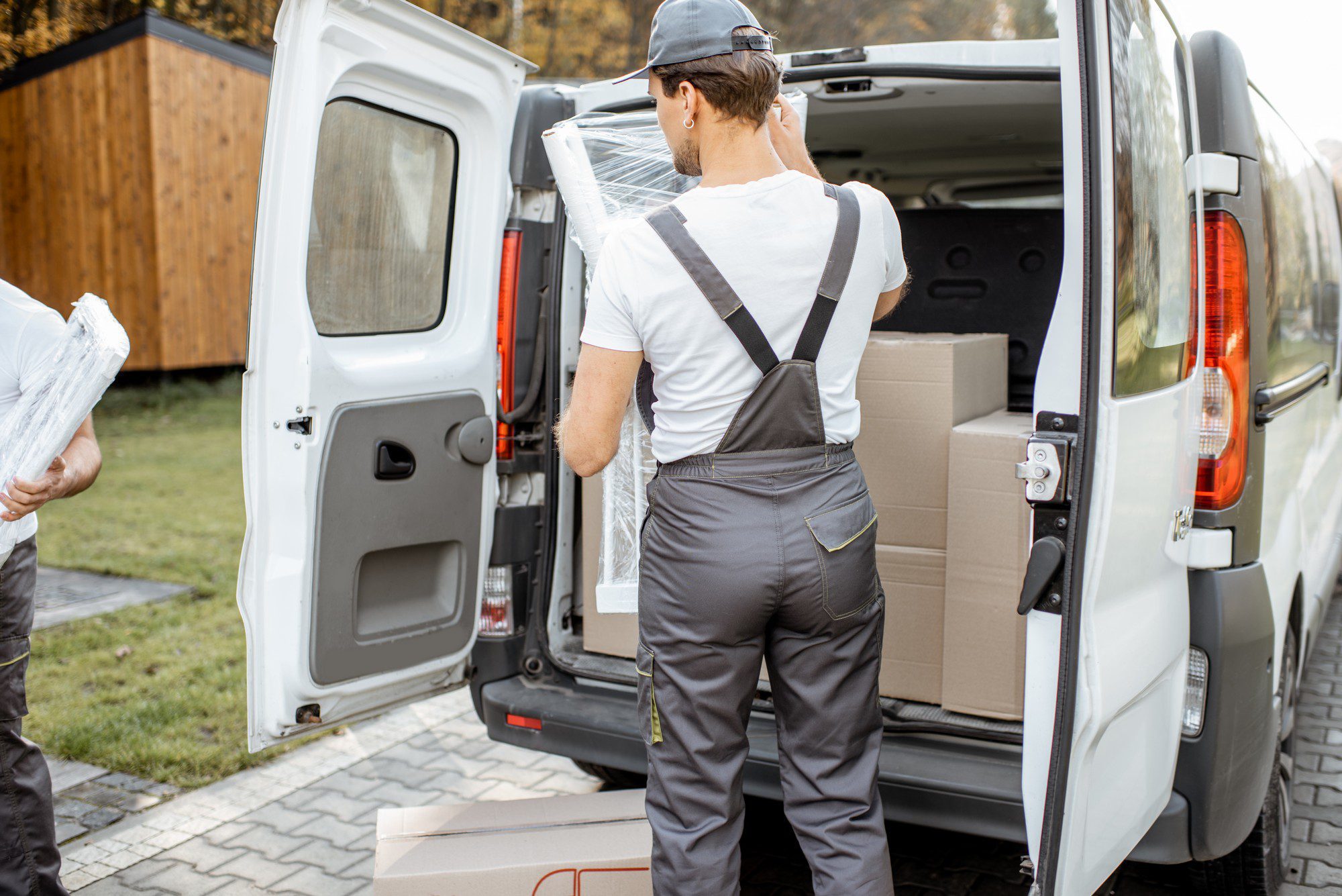 The image shows two individuals who appear to be in the process of loading or unloading a white commercial van. One person is mostly facing away from the camera, wearing a white t-shirt with braces over grey pants, and is reaching into the back of the van, which is open. Inside the van, there are cardboard boxes suggesting some kind of delivery or moving scenario. The second person, partially visible on the left side of the image, is carrying plastic-wrapped materials, which may be padding or protective wrapping for transporting fragile items. The setting seems to be a residential area with a paved driveway and some greenery in the background.