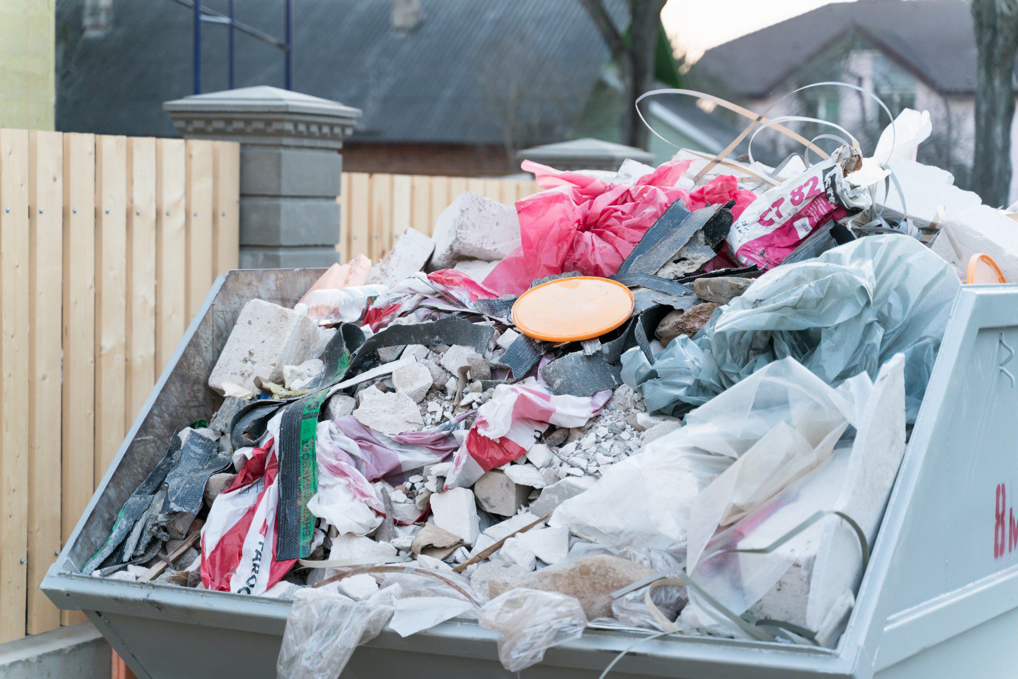 This is an image of a large metal skip or dumpster filled with various types of waste. It looks like there may have been a construction or renovation project, as the container is filled with debris such as broken pieces of concrete or plaster, parts of building materials, bits of wood, and plastic sheeting. There are also traces of other waste like a plastic bucket lid and some fabric or plastic bags. In the background, there is a wooden fence and a glimpse of residential buildings, indicating that this dumpster is likely placed in a residential area.