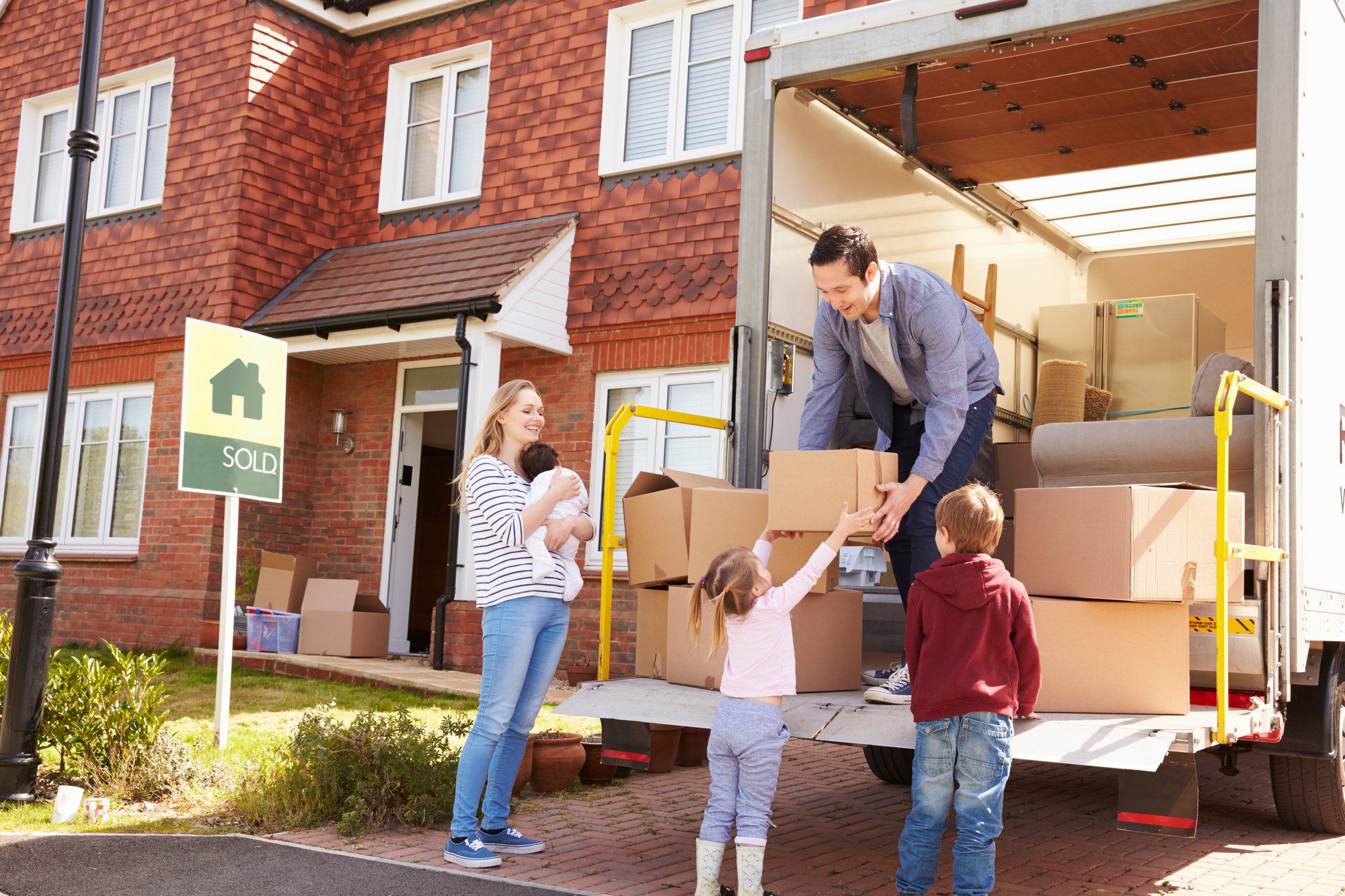 The image shows a family engaged in the process of moving into a new home. There is a 'SOLD' sign in front of the house indicating that the property has been recently purchased. A woman holding a baby is standing near the open door of the house, smiling and watching as a man hands a cardboard box to a young girl who is reaching up to take it. There is also a boy who appears to be waiting for a box or helping with the moving process. In the background, there is an open moving lorry loaded with furniture and boxes, suggesting that the family is unloading their belongings from the lorry into their new home. The scene is set in a sunny residential area, and the family looks happy and engaged in the experience.