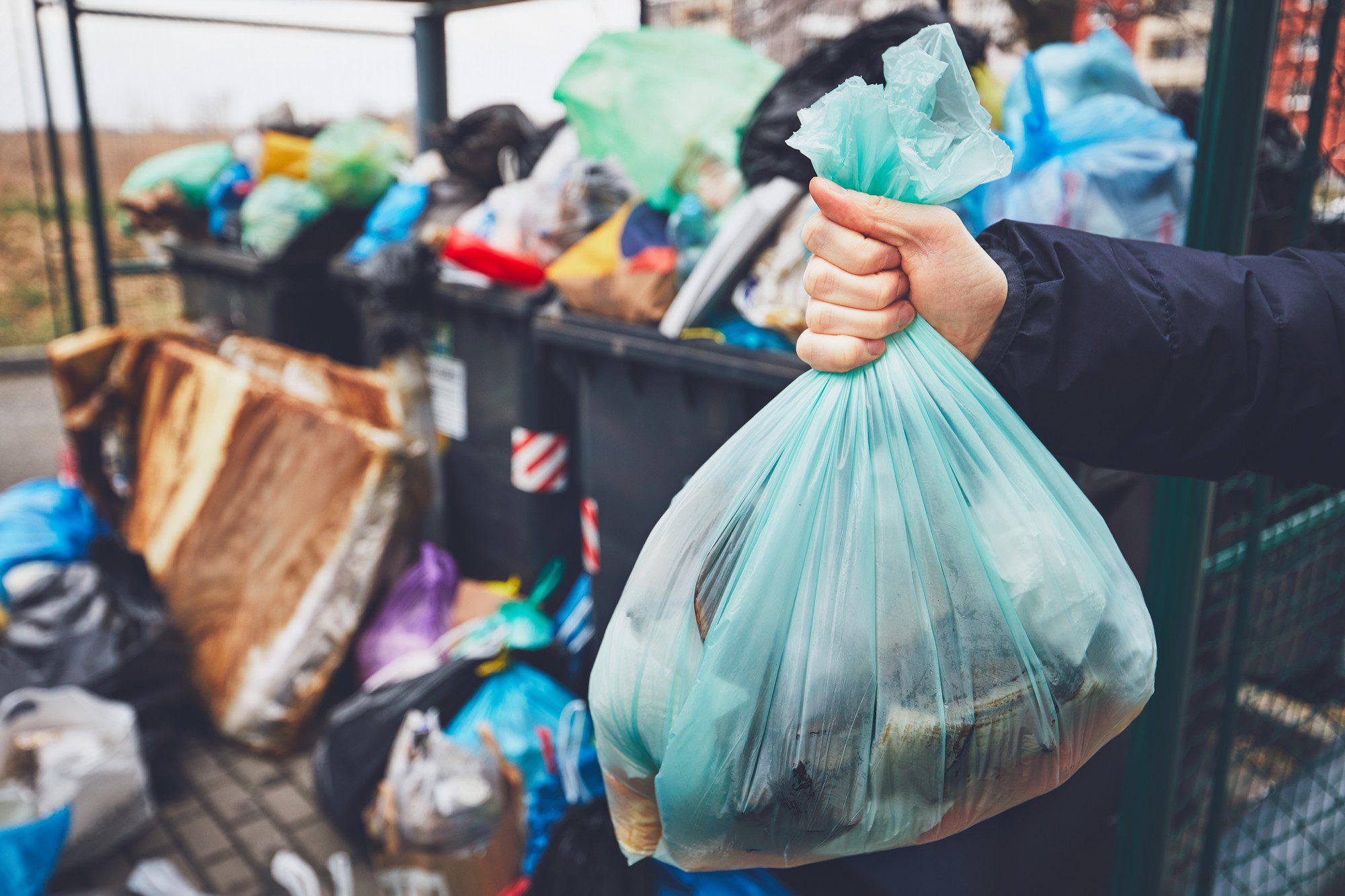 The image shows an individual holding a light blue plastic rubbish bag, which appears to be full. In the background, there are overflowing bins and more rubbish bags, suggesting that this is an area for waste collection that is quite full. The person is wearing a dark jacket, and only their gloved hand can be seen. The focus is on the hand holding the rubbish bag, with the bins and additional waste appearing out of focus in the background. This scene presents a common situation faced in urban areas where waste management facilities are sometimes insufficient for the amount of trash generated.