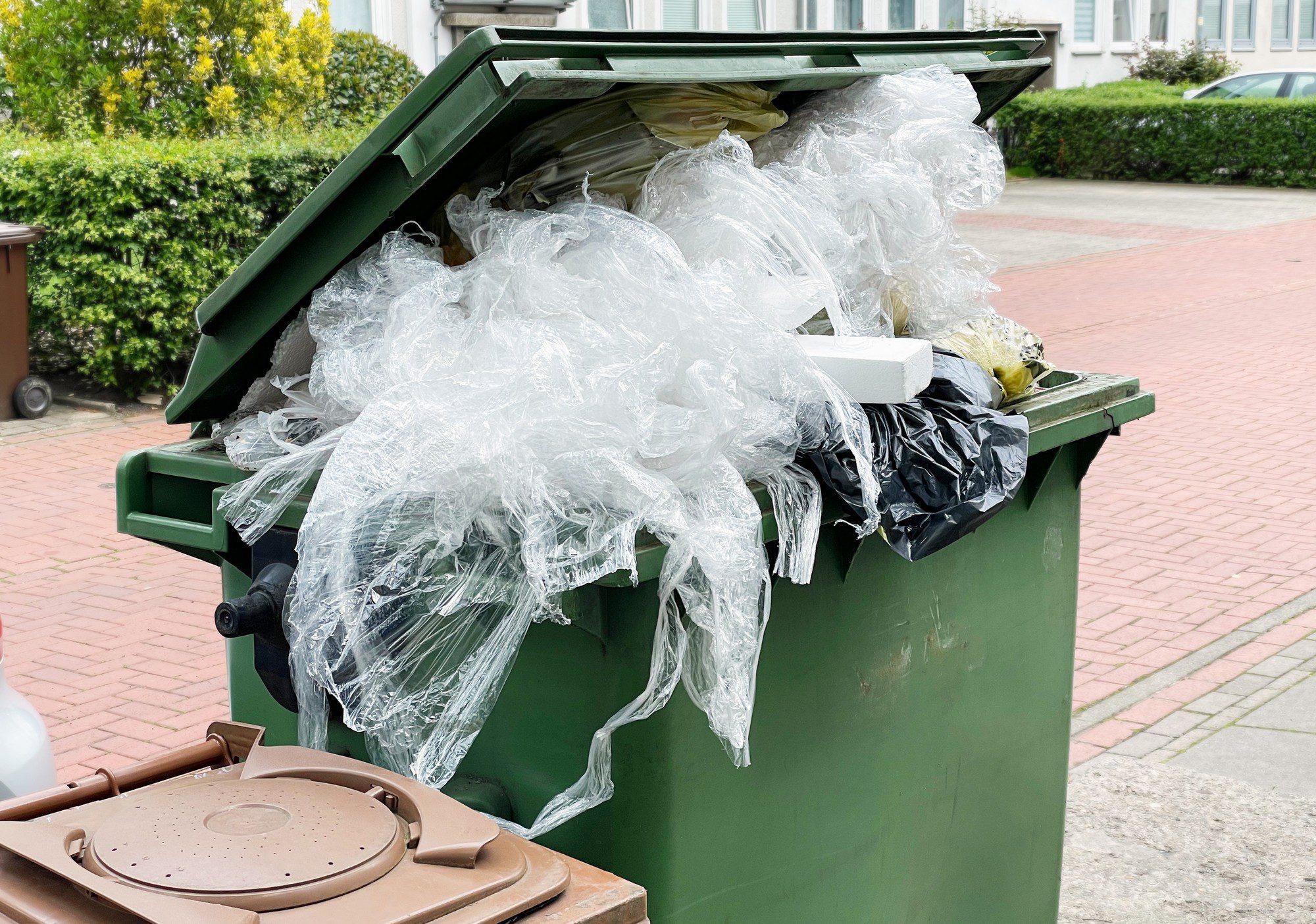 The image displays an overflowing green wheelie bin. The lid is ajar due to the excessive amount of waste stuffed inside, making it impossible to close properly. A large quantity of plastic film or plastic wrapping material spills out of the bin, and there are also visible pieces of foam or polystyrene, as well as a black plastic bag that also seems to contain waste. The environment around the bin looks like a residential area with paved ground and plants in the background. This image may bring attention to issues such as waste management, recycling, and the environmental impact of plastic pollution.