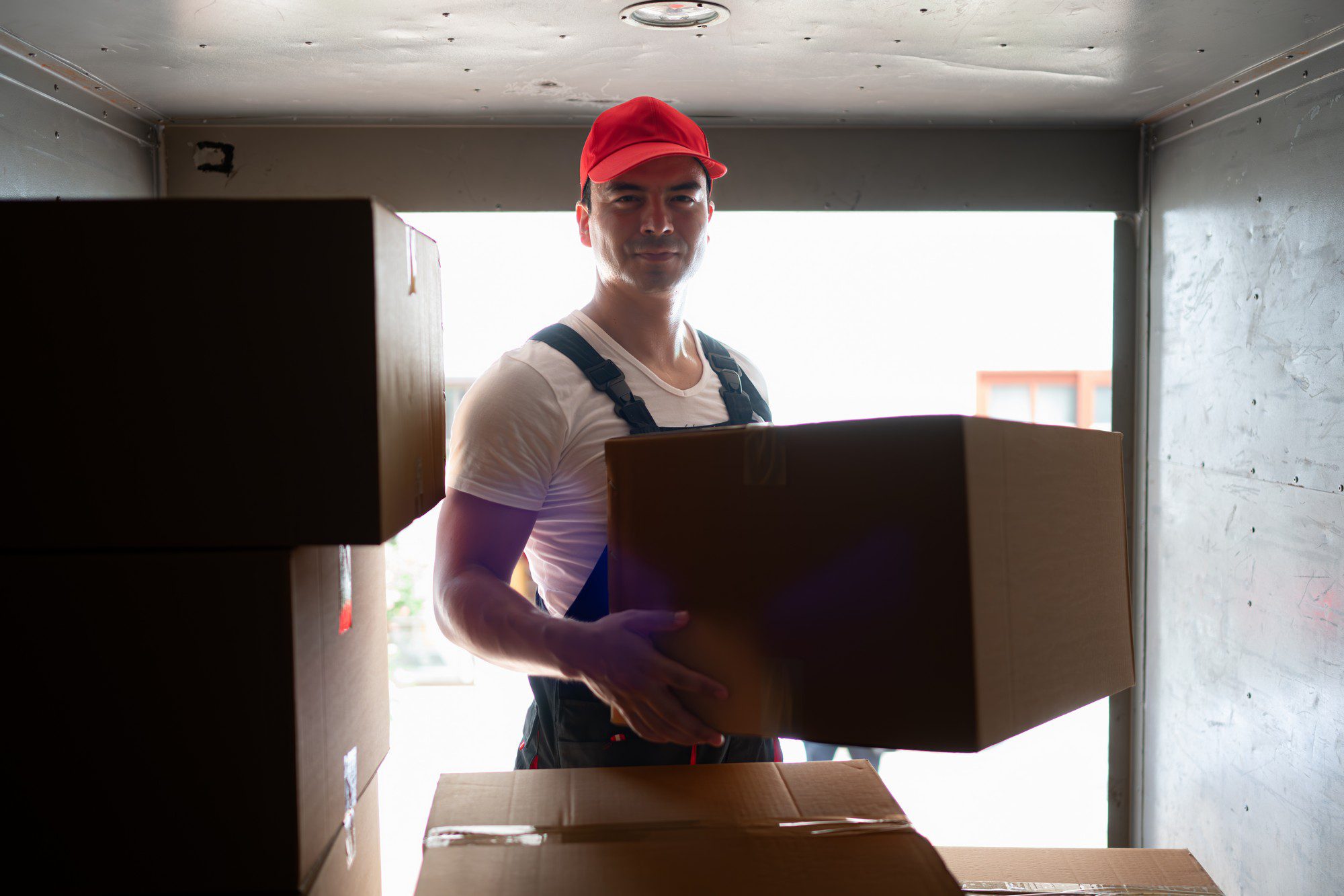 In the image, you see a person who appears to be a delivery man or mover. This individual is wearing a white t-shirt, a red cap, and blue braces over his shoulders, which may be part of a harness designed for lifting. He is holding a large cardboard box and appears to be standing inside a lorry or a storage area, where other boxes are stored. There's natural light coming from what seems to be an opening behind him, possibly the back of the lorry, suggesting that he might be in the process of loading or unloading items. The person is looking directly at the camera with a neutral to mild smile on his face.