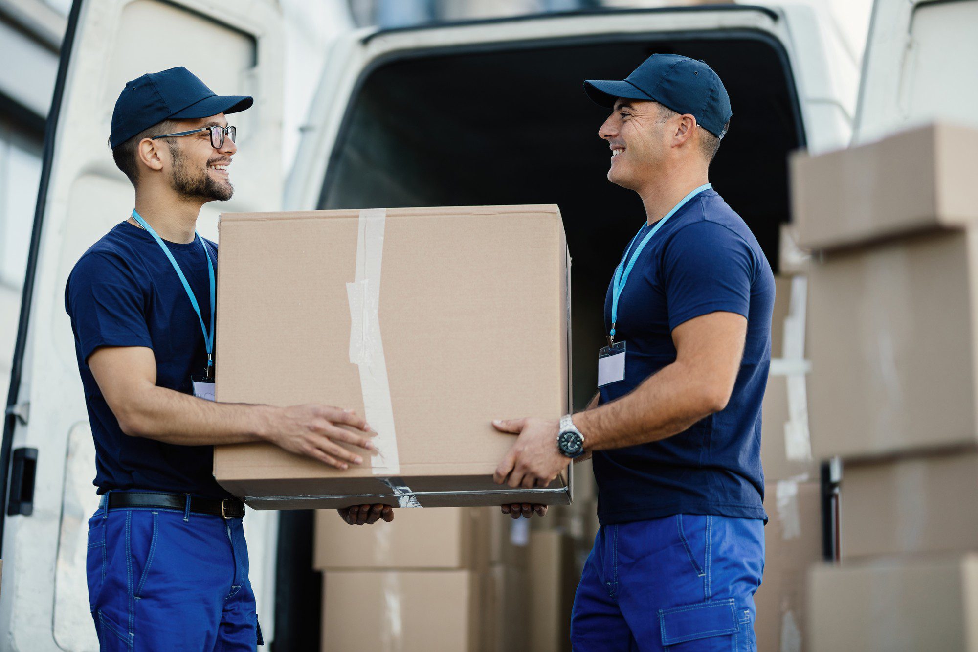 The image shows two individuals dressed in similar blue uniforms, including caps and a name badge or ID tag, handling a large cardboard box. They both appear to be delivery workers or movers, and they are standing in front of an open cargo van or delivery lorry, which is partially visible in the background. The workers are smiling and looking at each other, indicating a positive interaction, possibly while engaged in the process of loading or unloading the box from the vehicle. There are also additional cardboard boxes visible behind them, suggesting that there might be more items to deliver or move.