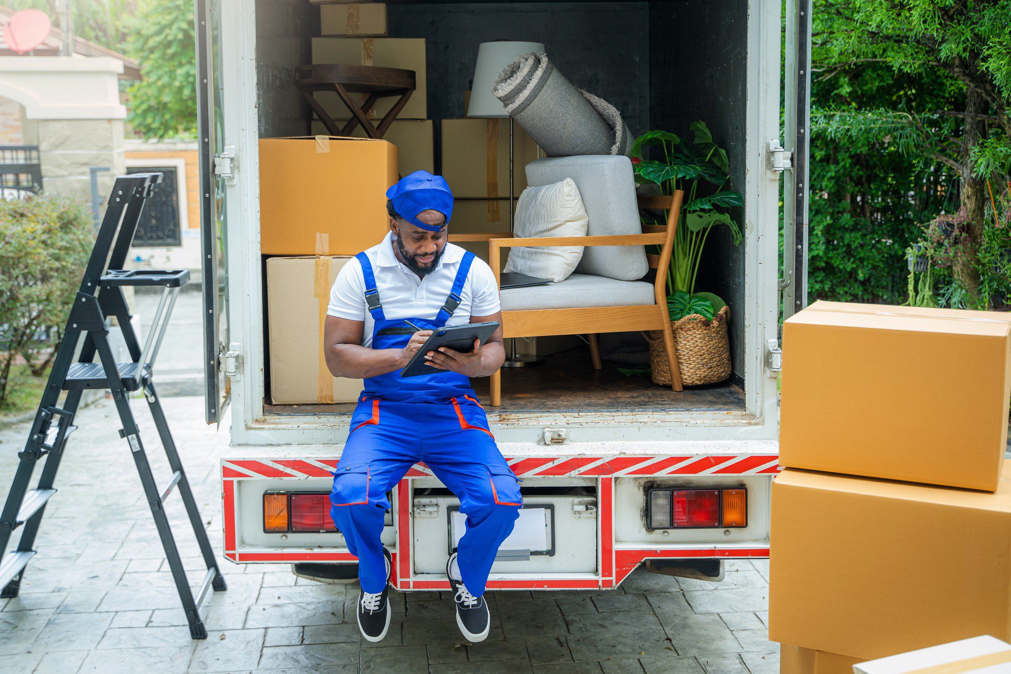 The image shows a man wearing a blue uniform with white and orange trim, who appears to be a mover. He is sitting on the loading ramp of a moving lorry, looking at a tablet. In the lorry, there are various items suggesting household goods in transit, such as a couch with pillows, a rolled-up carpet, boxes, and a step ladder. There are also cardboard boxes outside the lorry, indicating that the moving process is either in progress or nearly complete. The setting looks residential with greenery in the background, which indicates that the location could be a house or flat complex.