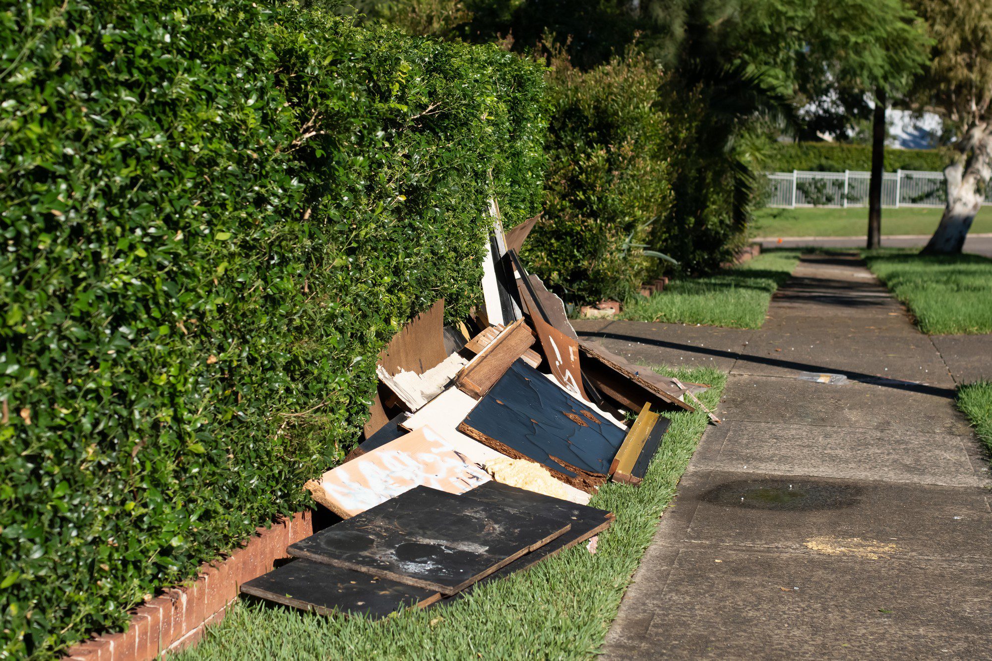 The image shows a collection of discarded items on the side of a residential sidewalk. There is a variety of debris that appears to be parts of wooden furniture or similar materials, such as boards and panels, some of which have paint or laminate surfaces. The debris is scattered and leaning against a trimmed hedge that lines the sidewalk, indicating it may be set out for bulk waste pickup.

The sidewalk runs parallel to a street with a grassy verge separating them. It's a clear day, and the shadows indicate it might be morning or evening due to the angle of the sunlight. There are trees and a fence visible in the background, which contribute to the suburban feel of the scene. The surrounding area appears well-maintained, suggesting that the debris placement is temporary, likely for disposal.