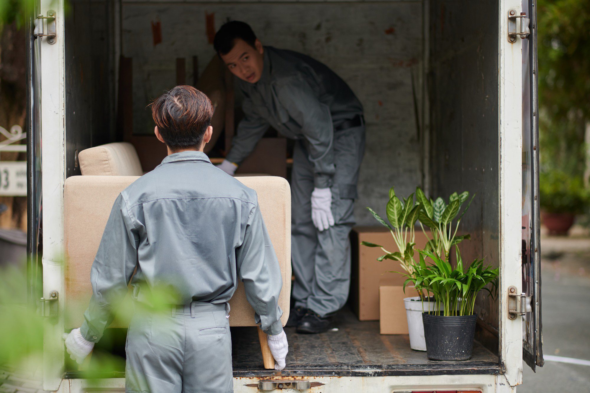 The image shows two individuals dressed in work uniforms involved in moving items. The person in the foreground has their back to the camera and is lifting one end of a beige armchair onto the back of a lorry, while the person inside the lorry is preparing to receive the armchair. There are also a few potted plants and cardboard boxes visible on the back of the lorry, indicating additional items being moved. The setting appears to be during daytime with an overcast sky, and the environment suggests they could be in a residential area. The whole scene is indicative of a moving process, where belongings are being transported from one place to another.