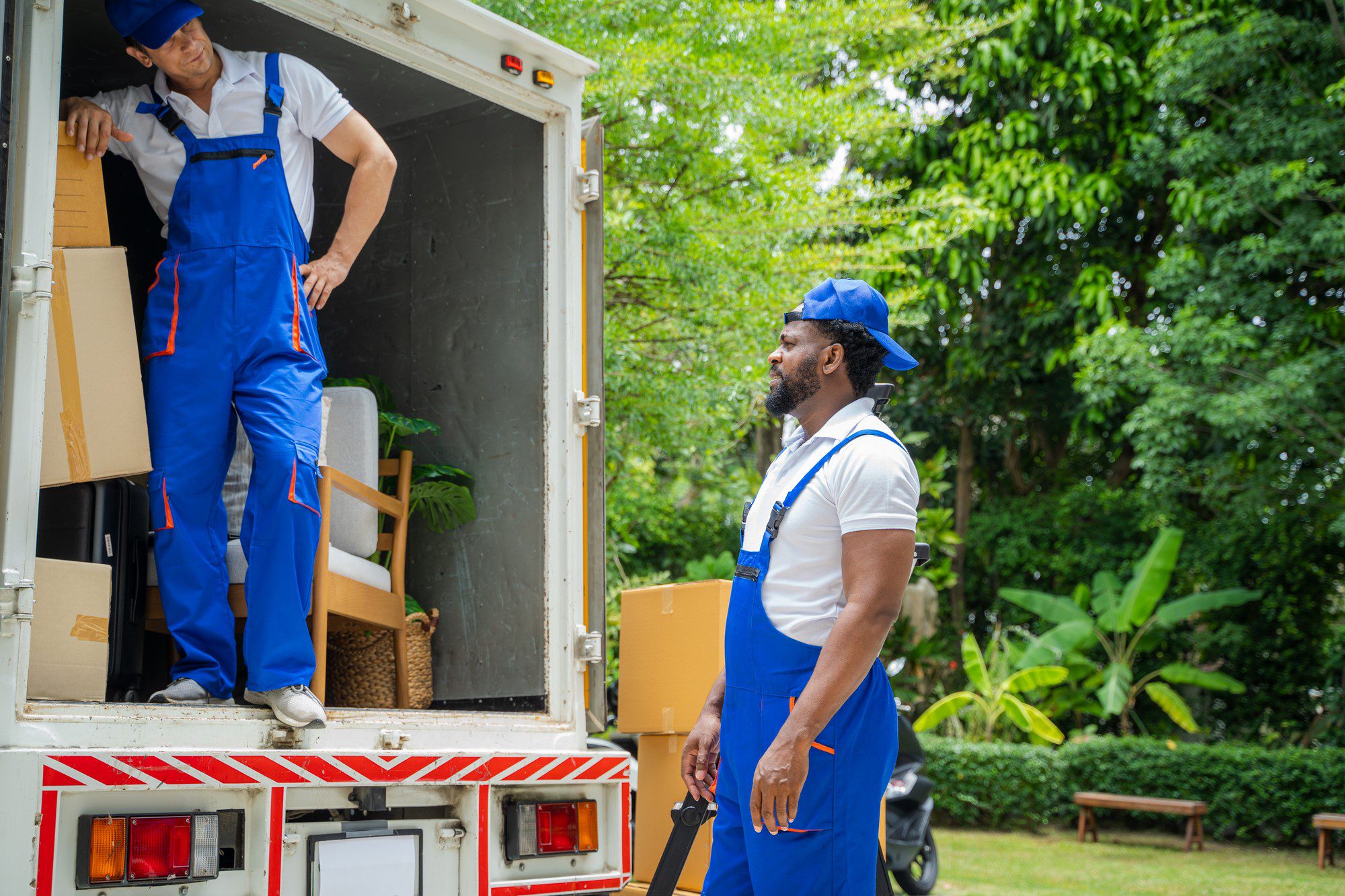 In the image, there are two individuals wearing blue uniforms with white undershirts, which suggest that they may be workers or movers. The one on the left is situated inside a white moving lorry and appears to be adjusting or moving the boxes and furniture inside, while the one on the right is outside the lorry, standing and holding what looks like a moving strap or harness. Surrounding them is a lush green environment with trees and plants, indicating that they are in a garden or park-like area. The rear door of the moving lorry is open, and several cardboard boxes and a piece of furniture are visible inside the lorry, suggesting they are either loading or unloading the vehicle as part of a moving process.