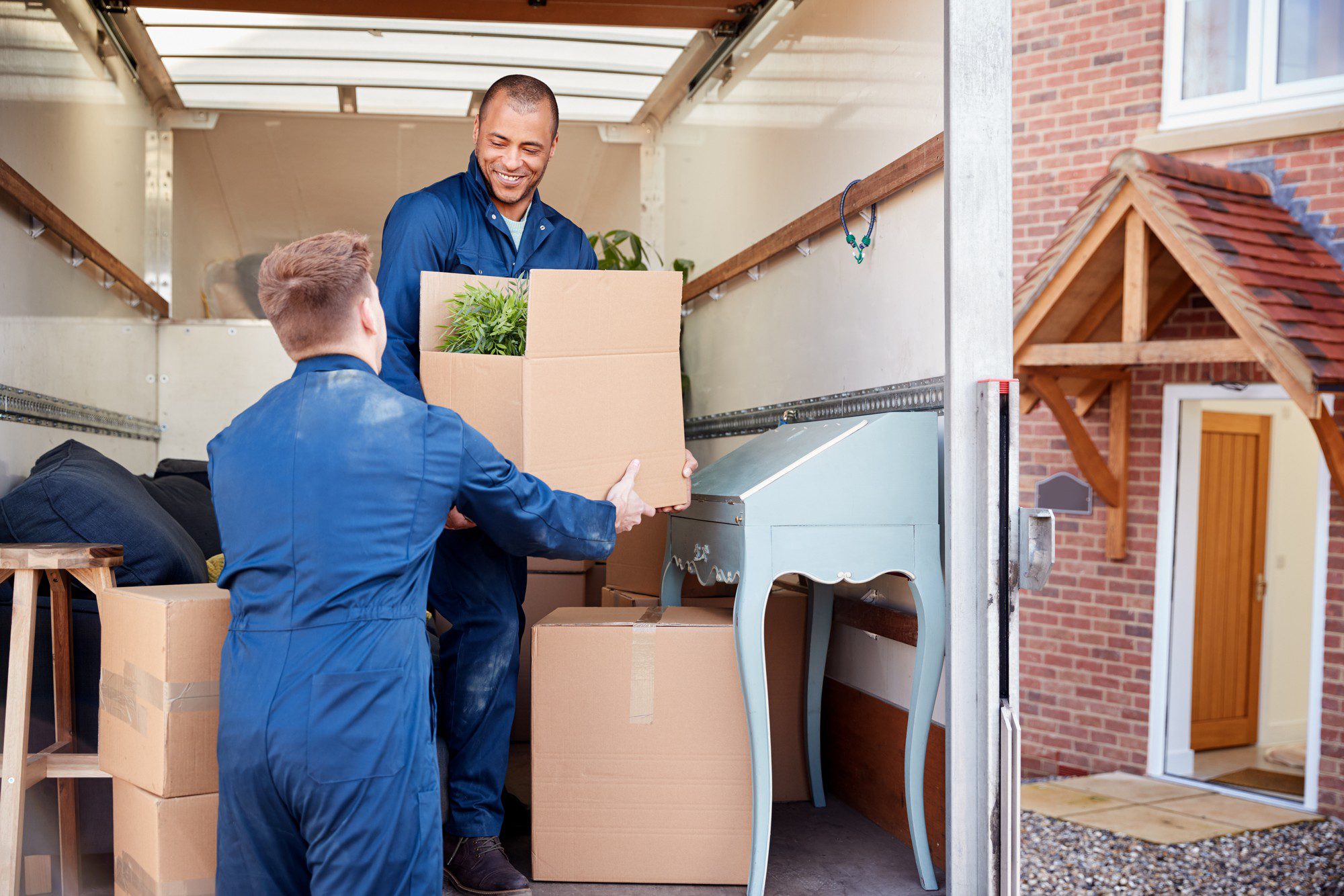 This image shows two individuals in blue work attire who appear to be moving items. They are handling cardboard boxes, one of which contains a plant, and they seem to be either loading or unloading these items from a lorry. In the background, you can see the exterior of a residential building, likely indicating that this is a scene from a moving day where belongings are being transported to or from a home. The person facing the camera is smiling, which gives the scene a positive and friendly atmosphere. There is also a piece of furniture, a light blue writing desk, partially visible inside the lorry. The environment suggests a clear day, possibly conducive to moving activities.