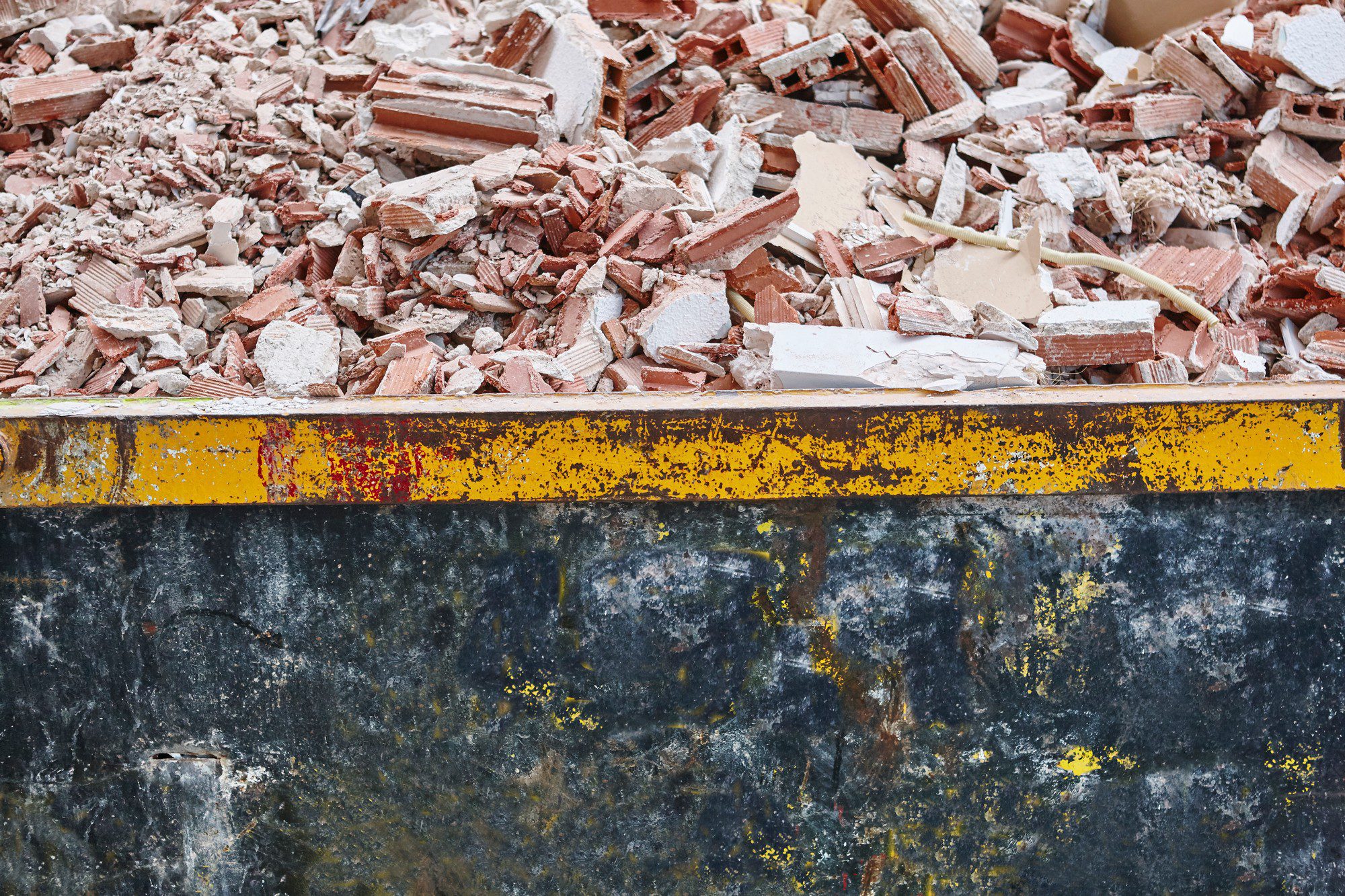 This image shows a large metal container, often referred to as a skip or dumpster, filled with construction debris. The debris primarily consists of broken pieces of bricks and what appears to be chunks of concrete or plaster. The edges of the container have visible yellow paint with signs of wear and rust, indicating that the container is used frequently and subjected to rough handling typically seen on construction sites. It's a scene that suggests demolition work or a significant renovation project where old building materials are being removed.