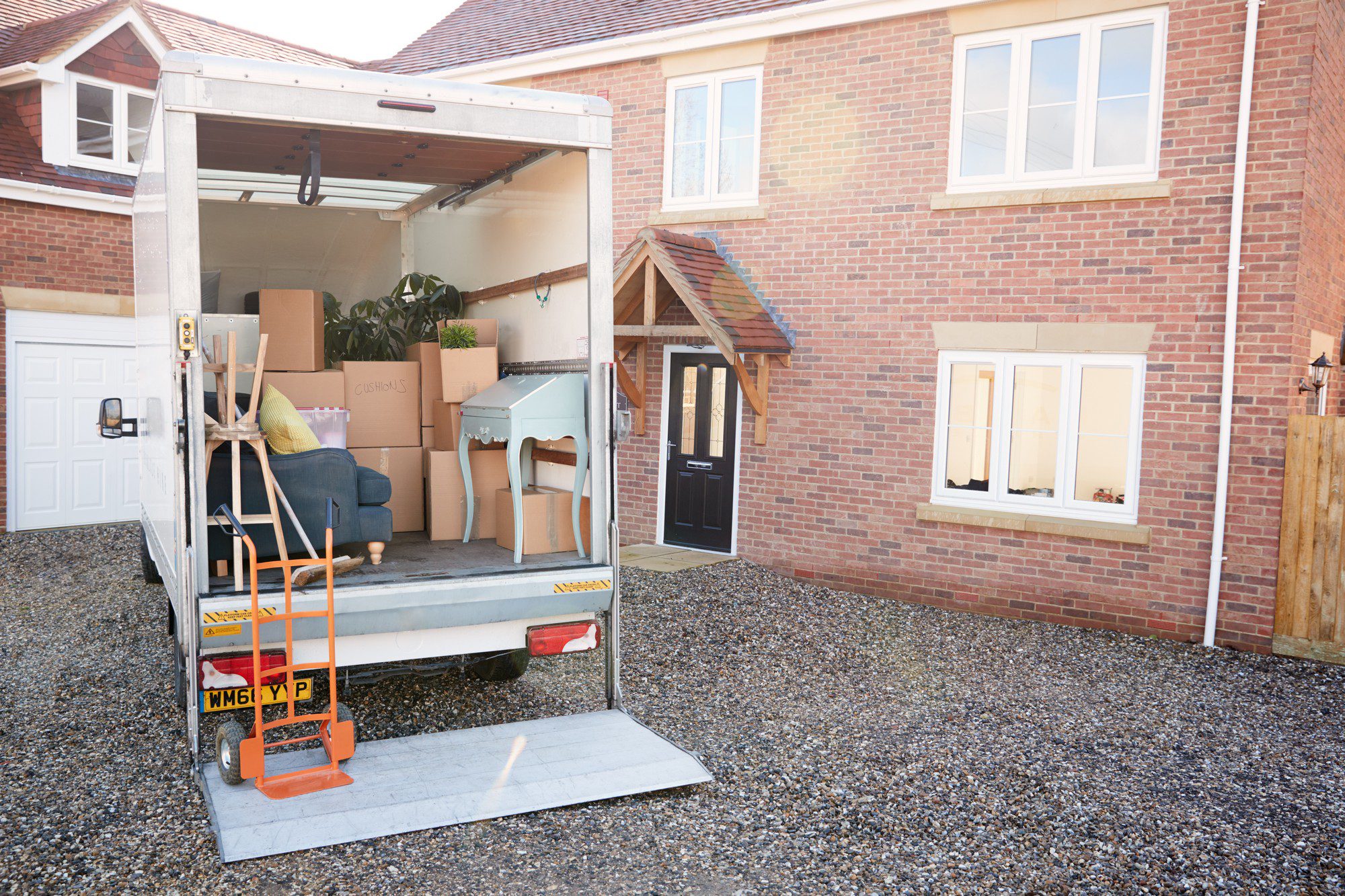 This image shows a moving lorry parked in front of a residential home. The rear gate of the lorry is open, and we can see various household items and cardboard boxes inside, indicating that someone is likely in the process of moving in or out of the house. Visible items include a blue upholstered sofa, a green plant, a wooden chair with a blue cushion, and numerous boxes that appear to be packed for moving. A hand lorry (dolly) can be seen at the edge of the lorry's lift gate, which is laid down to provide an easy way to load or unload the vehicle. The house is a two-story brick structure with white double hung windows and a black front door under a small covered porch with a gabled roof. A garage with a white door is also visible on the left side, and the driveway is composed of loose gravel or small stones.