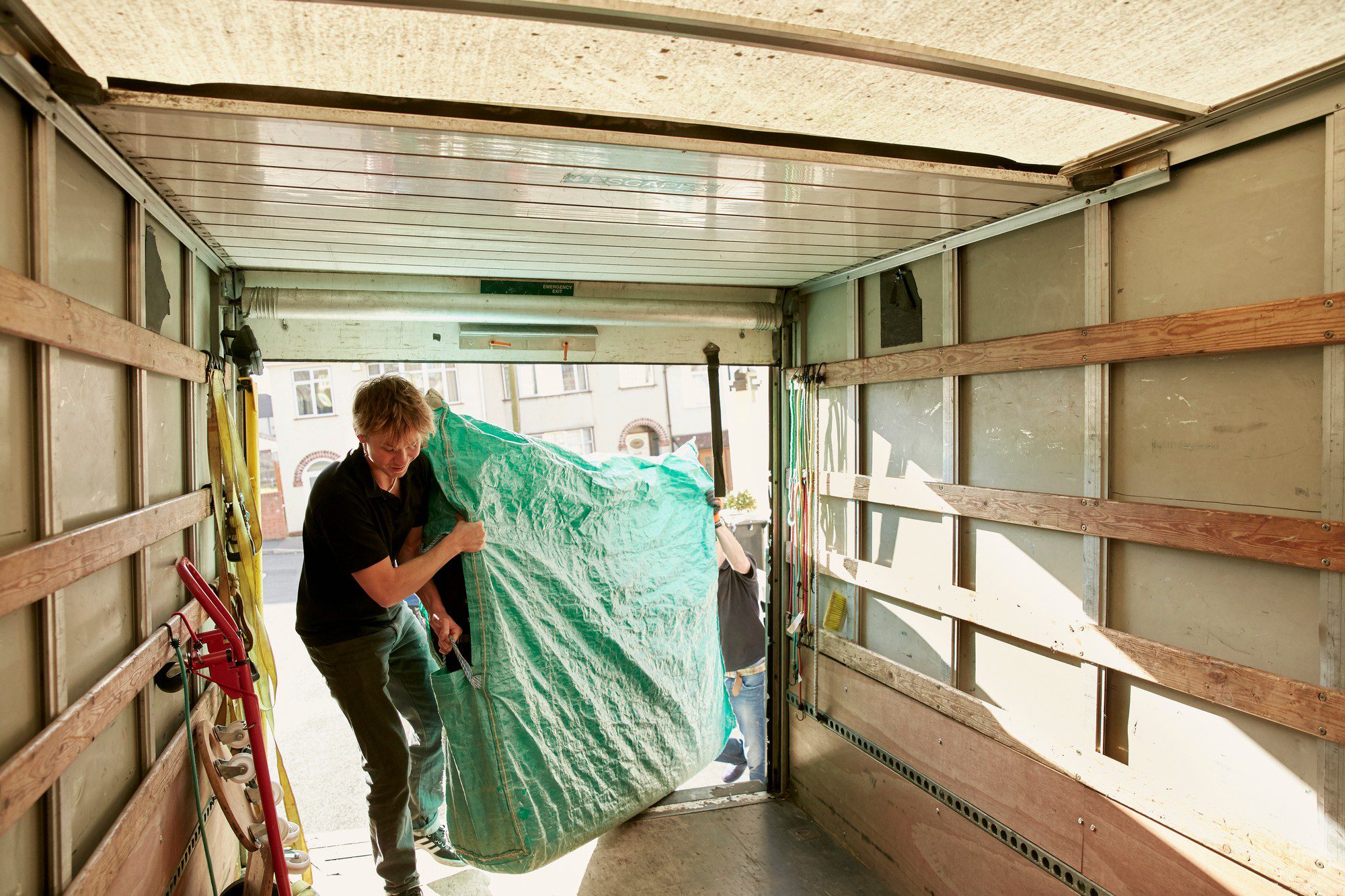 The image shows the interior of what appears to be a moving lorry, with the open back door revealing a view of a street and buildings outside. Two people are visible; one person is inside the lorry, facing away from the camera, handling a large, bulky item covered with a blue-green tarp. The other person is at the threshold of the lorry, primarily obscured by the tarp, assisting with the handling of the item, which could be a piece of furniture or another large object given the context of the image. You can see a variety of items and moving equipment inside the lorry, including a ladder, broom, and some straps hanging down the interior side of the lorry. The scene suggests the activity of moving items in or out of the lorry, typically associated with relocation or transportation of large objects.