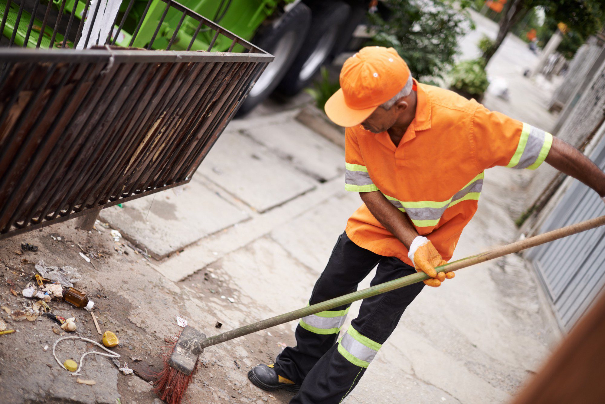 The image shows an individual wearing a high-visibility orange shirt and dark pants with reflective safety stripes, engaged in the task of sweeping a street or sidewalk. The person is using a broom with red bristles and a long wooden handle. There is litter on the ground, including pieces of paper, bottle caps, a glass bottle, and other debris near an iron fence. In the background, there appears to be a green vehicle and more people wearing similar high-visibility clothing, suggesting this is part of a coordinated cleanup or maintenance effort. The scene takes place outdoors during the daytime.