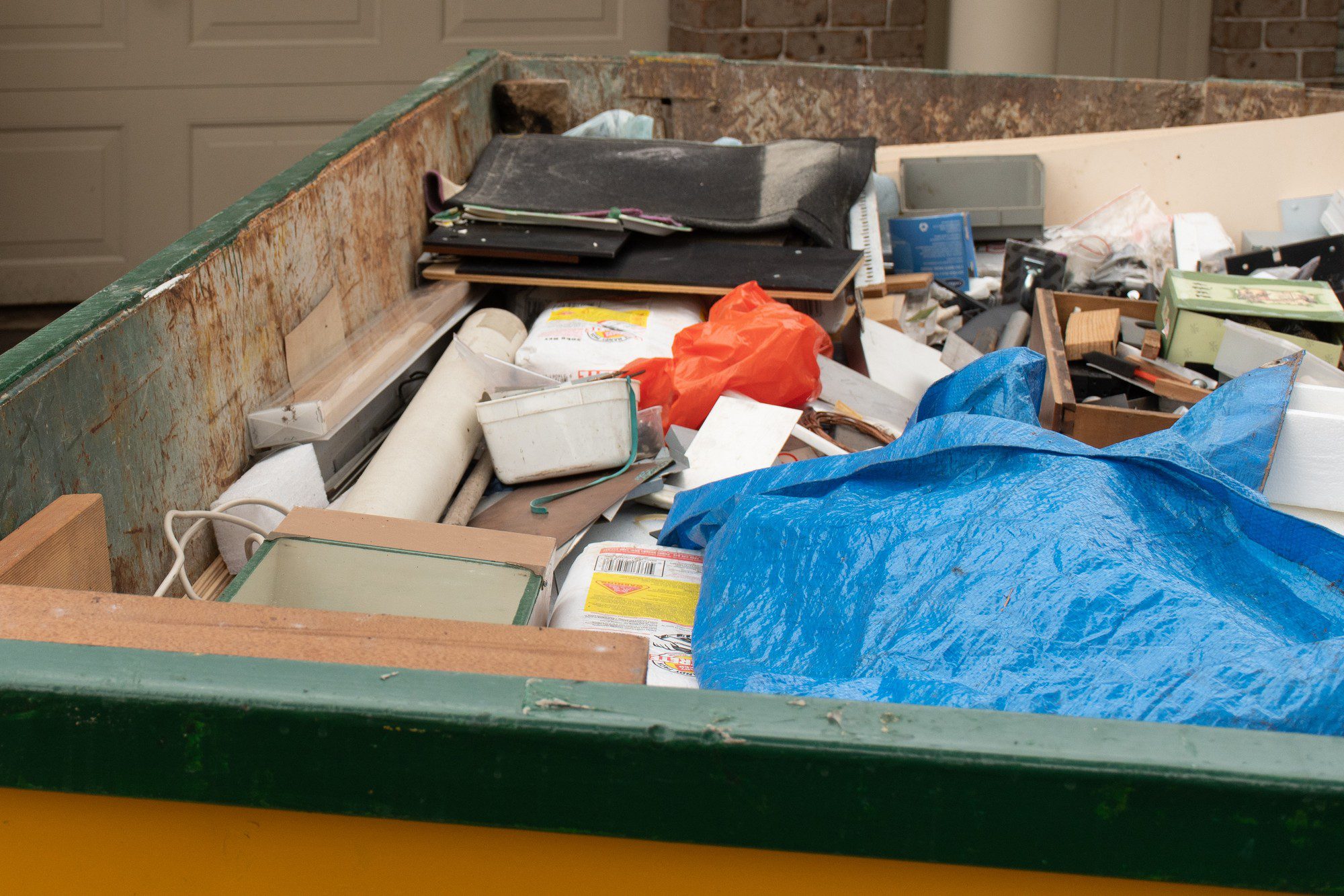 The image shows a large dumpster filled with various discarded items. Among the visible debris are an assortment of items that include:

- Building materials like rolled up rugs or mats
- Pieces of wood and wooden drawers
- Electrical items, possibly an old monitor or device
- Containers and plastic items
- Loose papers and cardboard
- A blue tarpaulin partially covering some of the items

The dumpster itself appears to be set in front of a residential garage door, suggesting it might be in use for a home cleanout, renovation, or construction project. The dumpster is rusted and shows signs of wear and age. The varied nature of the contents suggests a significant clean-up or disposal of a diverse range of household items.