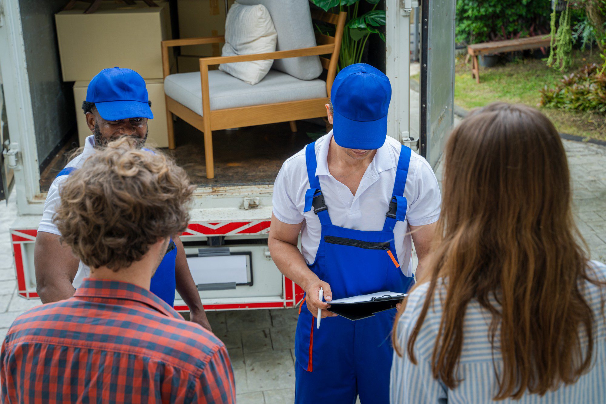 This image depicts a moving day scenario with professional movers and clients. In the foreground, we see two individuals, presumably the clients, with their backs to the camera. They appear to be conversing with a mover who is holding a clipboard, indicating they may be discussing details of the move, such as inventory, services, or paperwork.

The mover is wearing a white shirt with blue overalls, a blue cap, and is equipped with a lanyard around his neck, suggesting that he could be the moving team leader or supervisor. There is another mover in the background, visible inside the open moving lorry, seen through the rear doorway. He is also wearing a blue cap and overalls, indicating he is part of the moving team.

The moving lorry is loaded with items like boxes and furniture, with a couch and wrapped chair visible through the door. The setting seems to be a residential area, with plants and a lawn visible behind the lorry, contributing to the casual, suburban feel of the moving situation.