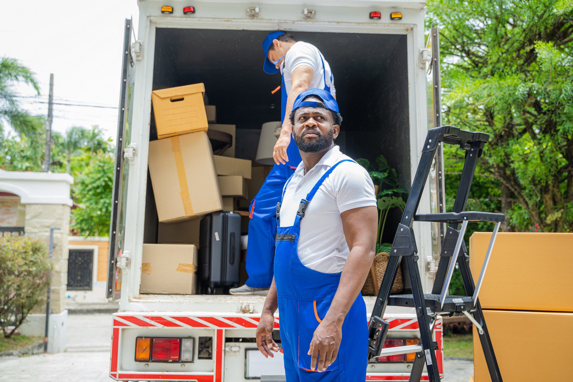 This is an image of two individuals who appear to be movers. They are wearing blue uniforms with white shirts underneath. The man in the foreground is standing outside of a moving lorry and looking towards the camera, while the other man is inside the lorry arranging or moving boxes. There are various packed cardboard boxes and pieces of furniture in the moving lorry, indicating that they are either in the process of moving items into the lorry for a move or perhaps unloading them at a destination. The setting seems to be a residential area with vegetation and parts of a house visible in the background.