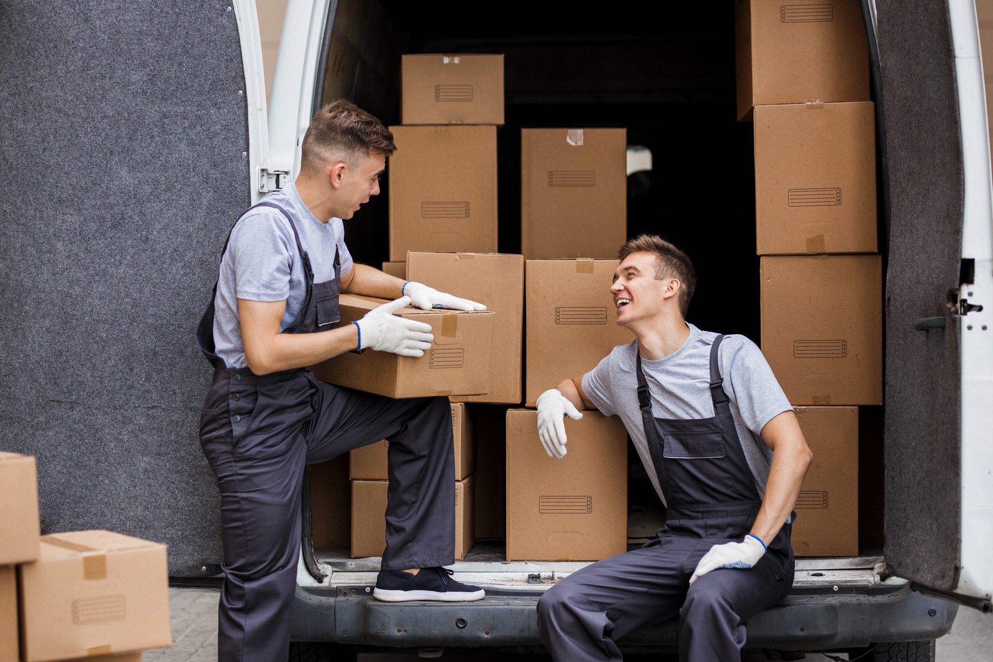 This image features two individuals who appear to be movers or delivery people, handling cardboard boxes near a white delivery van. They are wearing work uniforms, consisting of gray t-shirts and dark gray pants with protective gloves. One of them is sitting inside the van with boxes surrounding him, while the other stands at the van's rear, handing over a box. Both of them are looking at each other and seem to be engaging in a cheerful conversation or sharing a joke, as indicated by their smiles and body language. The setting indicates that they might be in the middle of loading or unloading the van.