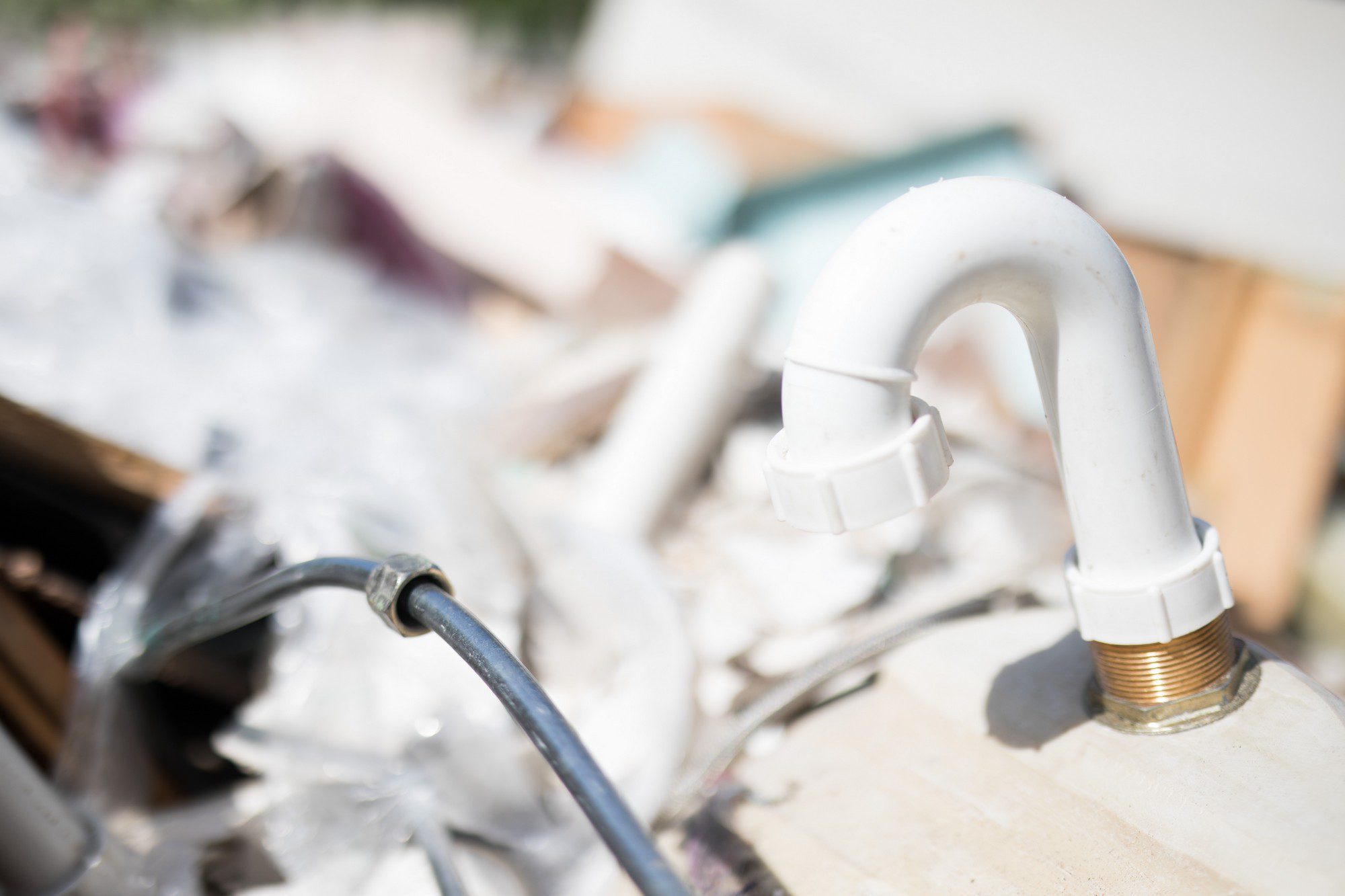 The image shows a close-up of a white U-shaped PVC pipe, which appears to be part of a sink's plumbing. This is typically the P-trap, a component that prevents sewer gases from entering the home through a drain. It holds a small amount of water which creates the seal. There is also a flexible metal tube in the bottom left portion of the frame, perhaps connected to a different part of the plumbing or some appliance.

The background is blurry, but it looks like there might be discarded items or debris, suggesting the setting could be a renovation site or a space where plumbing work is being done.