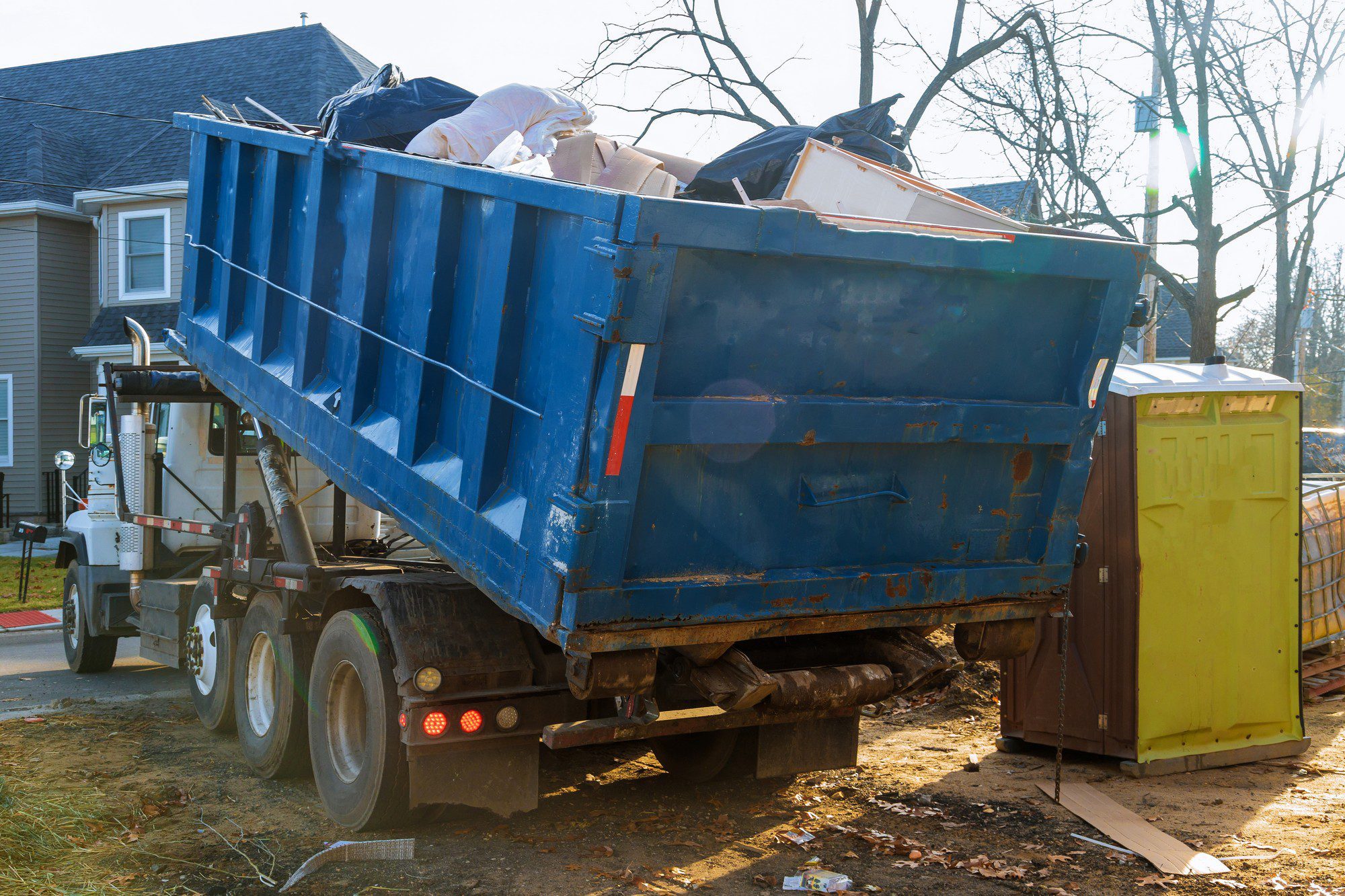 In this image, there is a blue roll-off dumpster on the back of a heavy-duty lorry. The dumpster is loaded with various discarded items including construction waste and debris. To the right side of the dumpster lorry is a portable toilet utility often used on construction sites or outdoor events. The background shows residential houses and trees, suggesting that the location could be a residential area, possibly undergoing construction or renovation work. The ground is somewhat muddy and scattered with small debris, implying recent activity in the area.