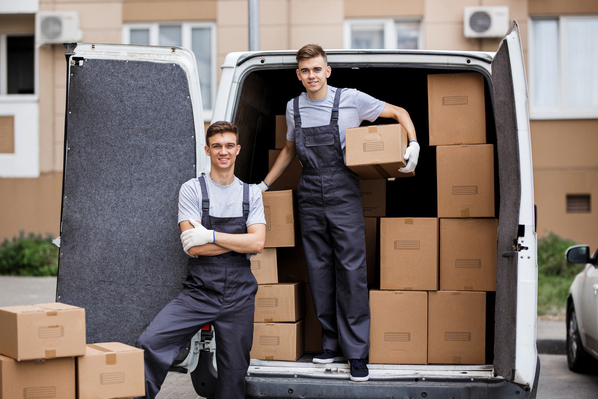 In this image, there are two individuals standing near a white van with its rear door open, revealing a stack of cardboard boxes inside. The people appear to be movers, based on their attire which includes matching dark overalls with light-coloured T-shirts underneath, and are wearing work gloves. Both individuals are young males, and one is holding a box, suggesting that they are in the process of loading or unloading the van for a moving job. The background indicates that they are in an urban environment, with flat buildings and a parked car visible. They seem to be smiling and looking at the camera, giving a positive impression of the moving process.